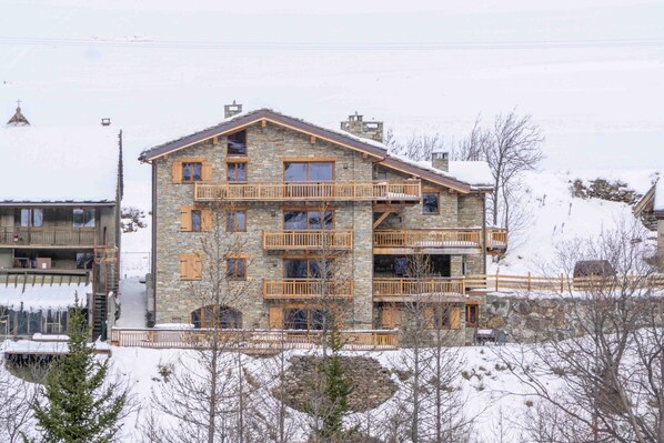 Un immeuble en pierre à plusieurs étages, doté de balcons, avec un télésiège en arrière-plan et de la neige au sol.