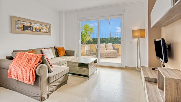 A living room with a sofa, coffee table, and TV. There is a sliding glass door leading to an outdoor area with a view of palm trees. - Holiday Apartment "Amanda" with Mountain View, Shared Pool & Wi-Fi (Xàbia)