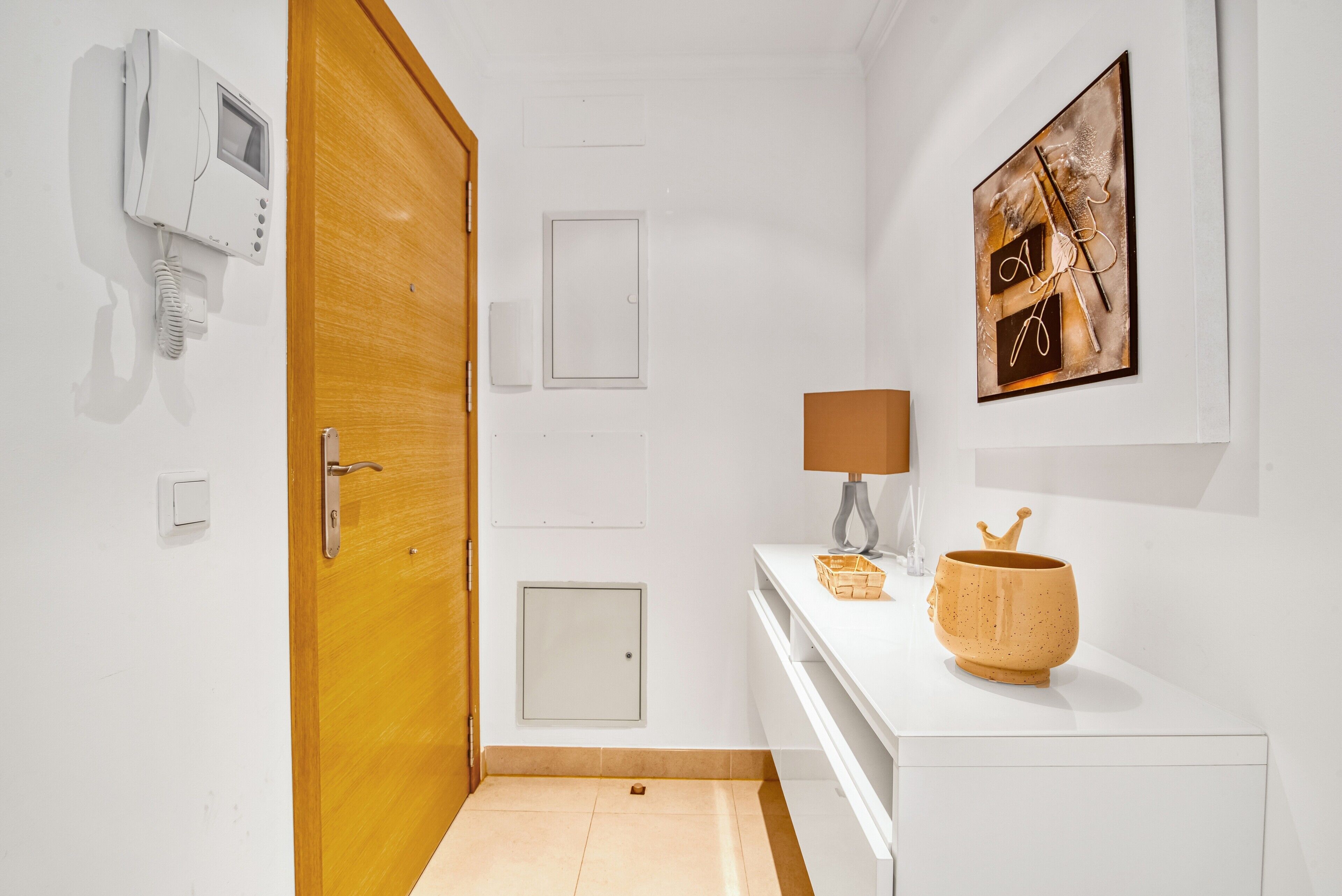 A hallway with a wooden door, a white wall-mounted phone, a small cabinet, a framed artwork, and a white counter with a lamp and decorative items.
