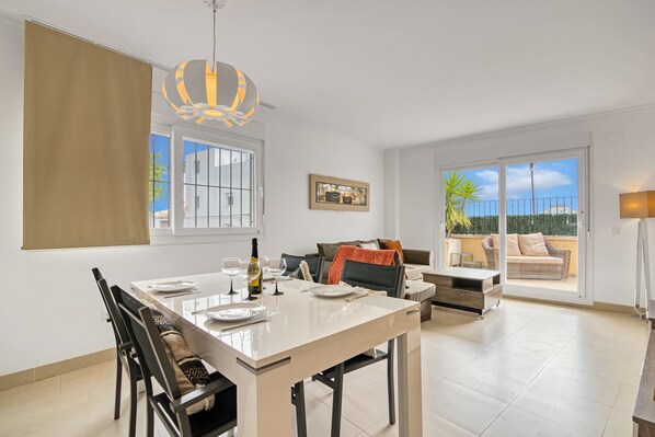 A dining area with a table, chairs, and a bottle of wine. There is a sofa and a window with a view of the outside.