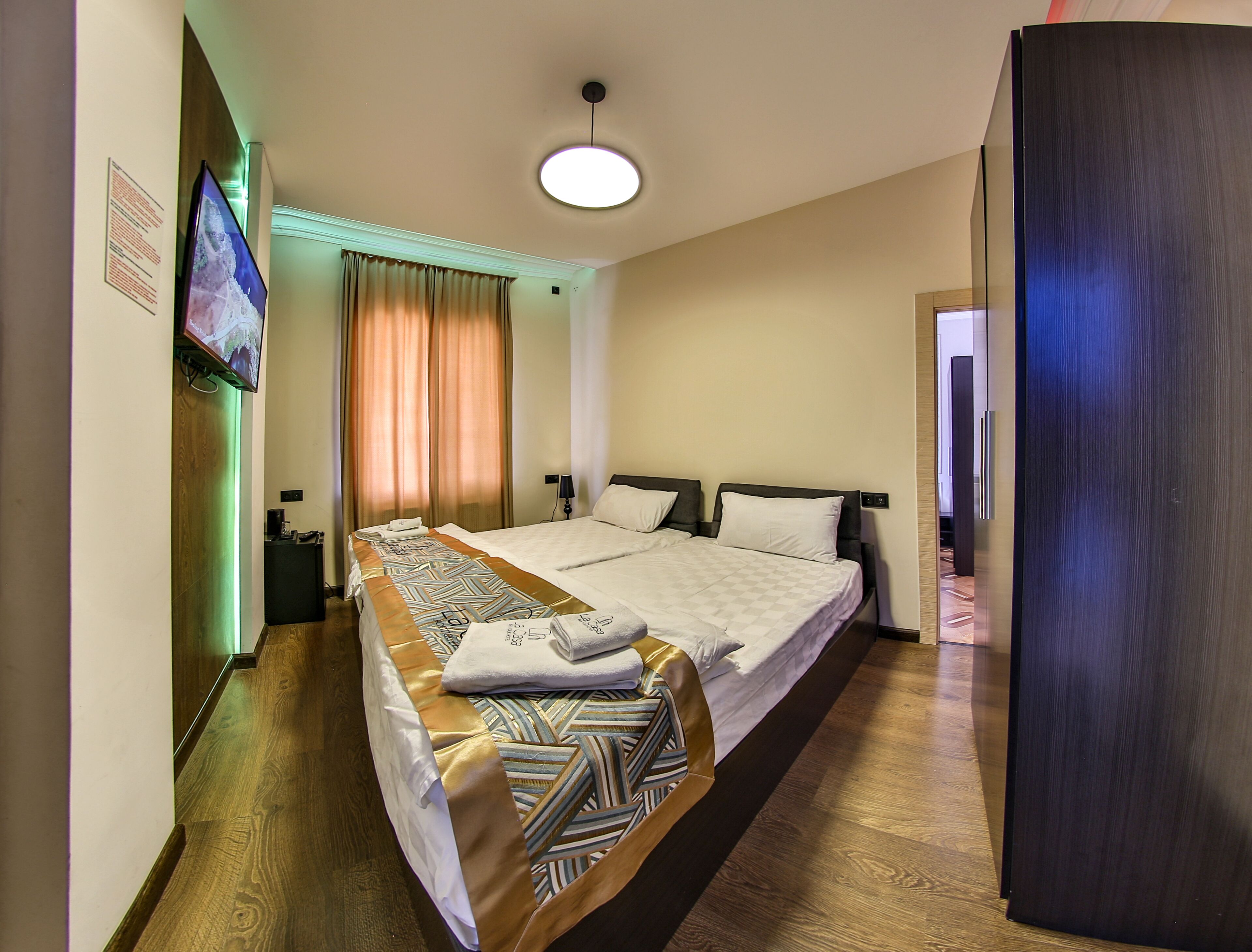 A hotel room with a large bed, a flat-screen TV mounted on the wall, a wooden floor, and a ceiling light.