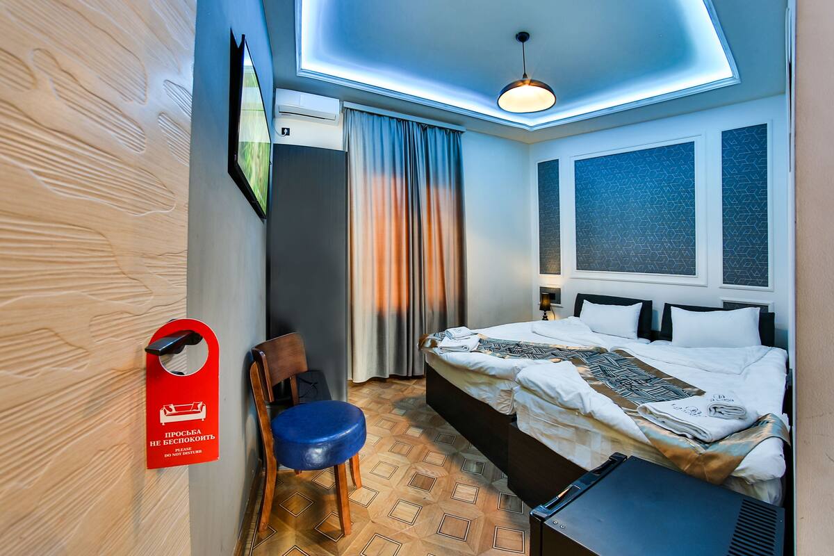 Ein Hotelzimmer mit Bett, Sessel, Schreibtisch, Deckenleuchte und einem fensterbepflanzten Fenster.