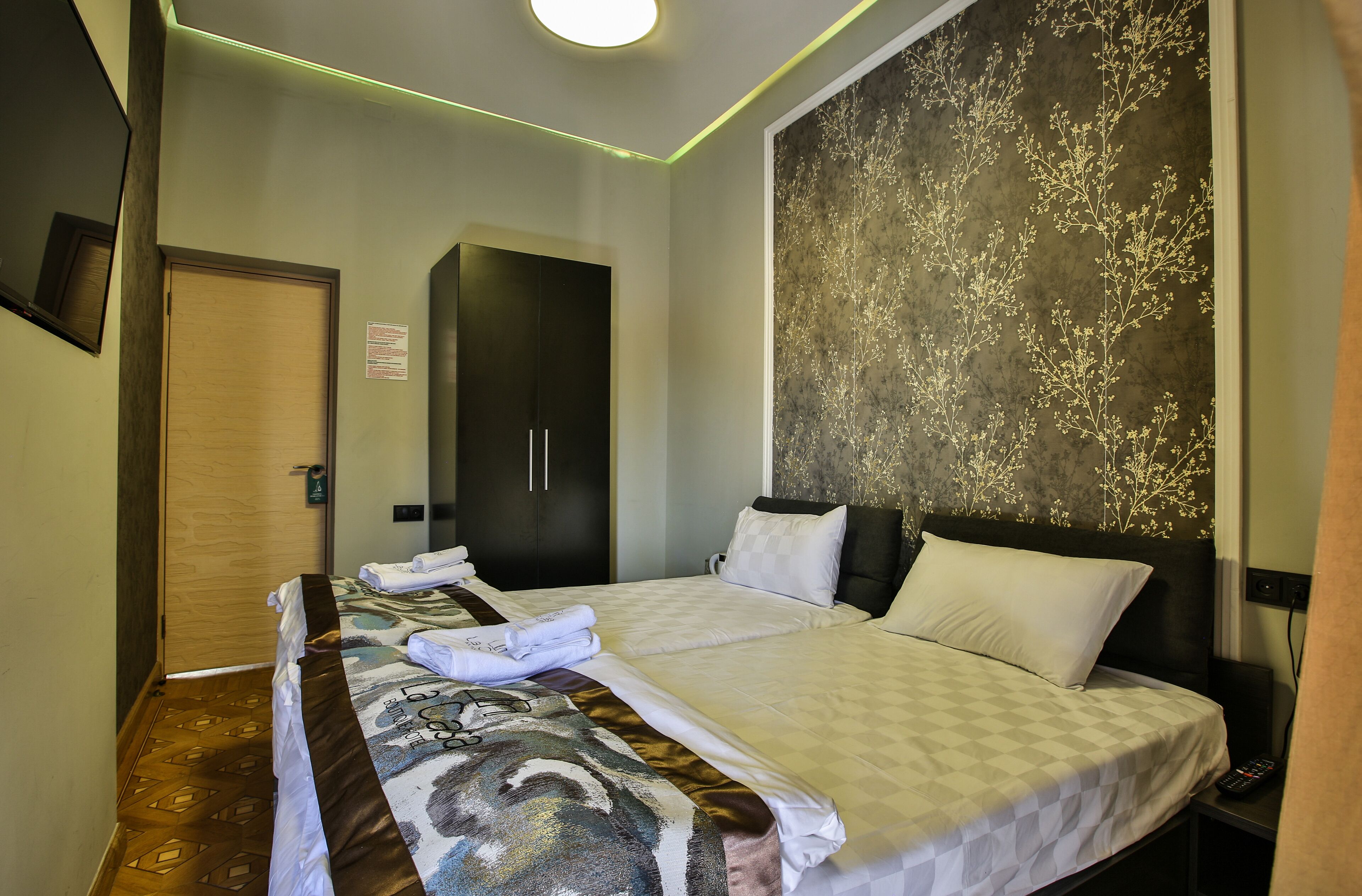A hotel room with a neatly made bed, a wardrobe, and a wall with a floral pattern.