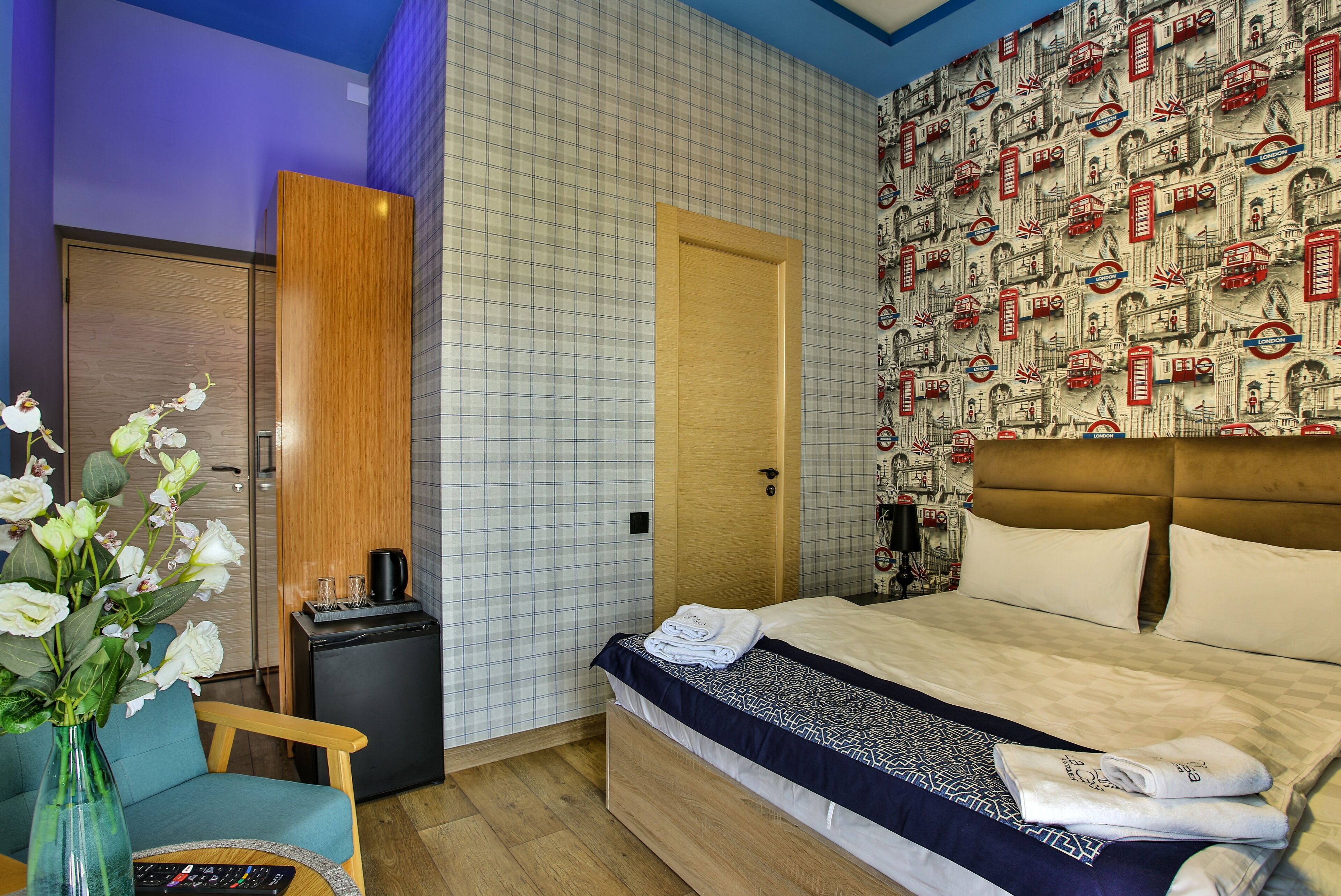 A hotel room with a bed, a small desk, a chair, a vase with flowers, and a wall with a checkered pattern.