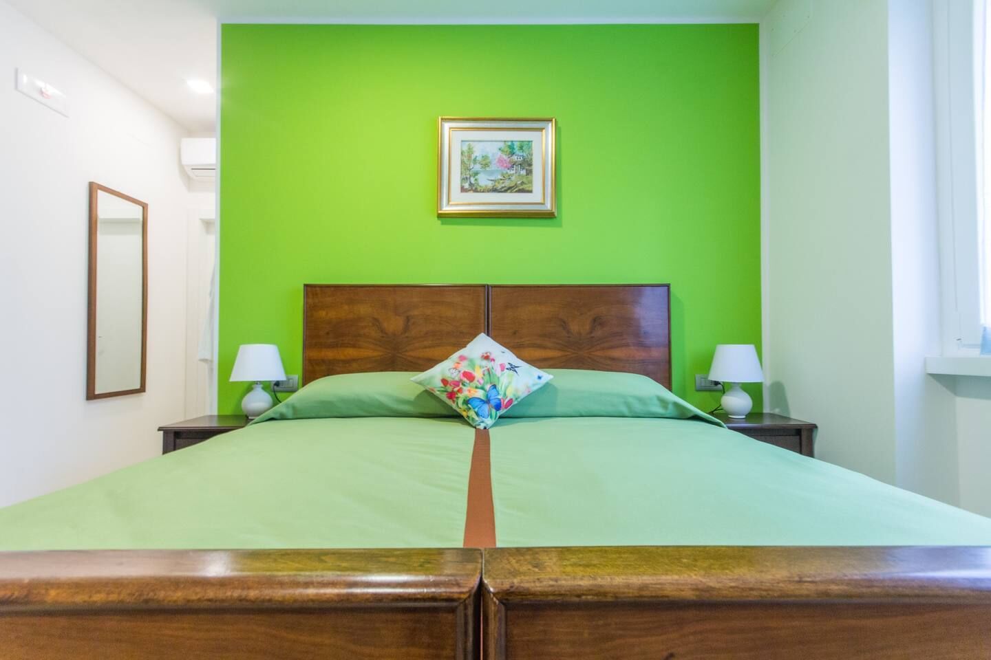 A bed with a wooden headboard, green bedding, and a floral pillow. There are two bedside tables with lamps, a framed picture on the wall, and a window on the right.