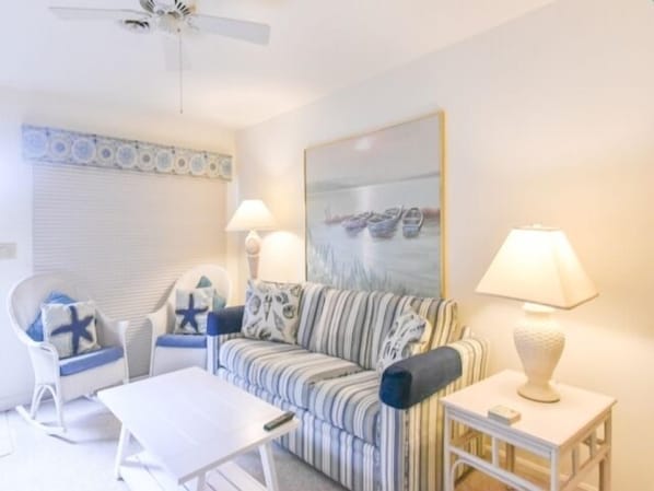 Una sala de estar con un sofá a rayas, un sillón blanco, una mesita y un ventilador de techo.