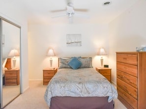 Un dormitorio con una cama, dos mesitas de noche con lámparas, un aparador de madera, un ventilador de techo y un espejo.