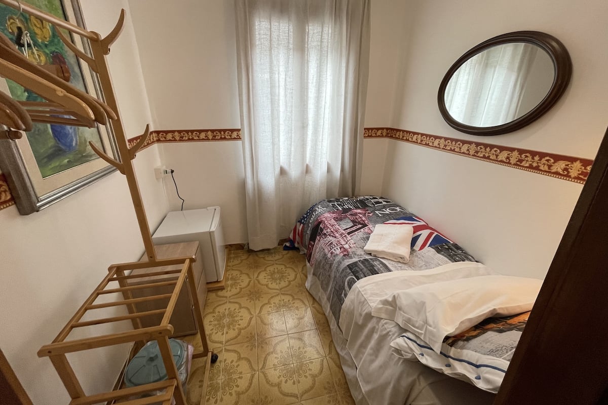 A small, compact room with a bed, a wooden chair, a small table, and a mirror on the wall.
