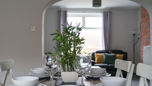 Un coin repas avec une table mise pour quatre personnes, comprenant des assiettes blanches, des bols et des verres à vin. Une plante en pot est placée au centre.