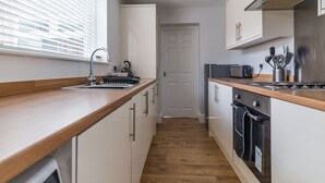 Une cuisine étroite avec un plan de travail en bois, des placards blancs, une cuisinière et une fenêtre avec des stores.