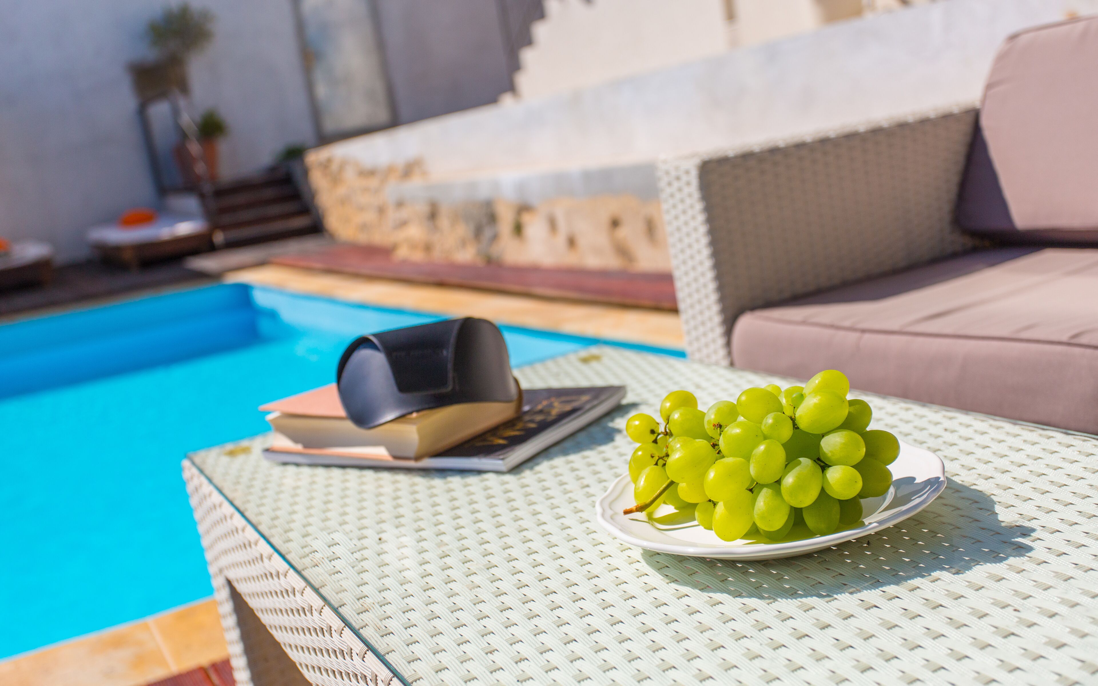 A poolside setting with a white woven table, a plate of green grapes, a book, and a black object on the table.