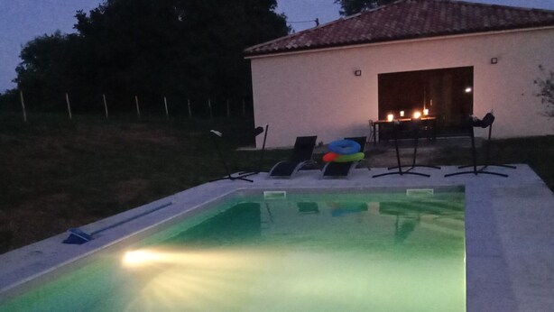 En swimmingpool med fliseterrasse, en liggestol og en lille patio med bord og stole.