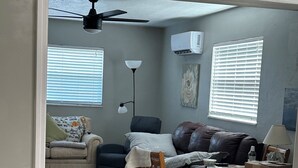 A living room with a ceiling fan, two windows with blinds, a couch, a chair, a table with items, and a floor lamp. - Beautiful, spacious one bedroom apartment. (Clearwater)