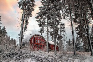 A red building surrounded by tall, snow-covered trees. - Hotel Obří Sud (Dlouhý Most)