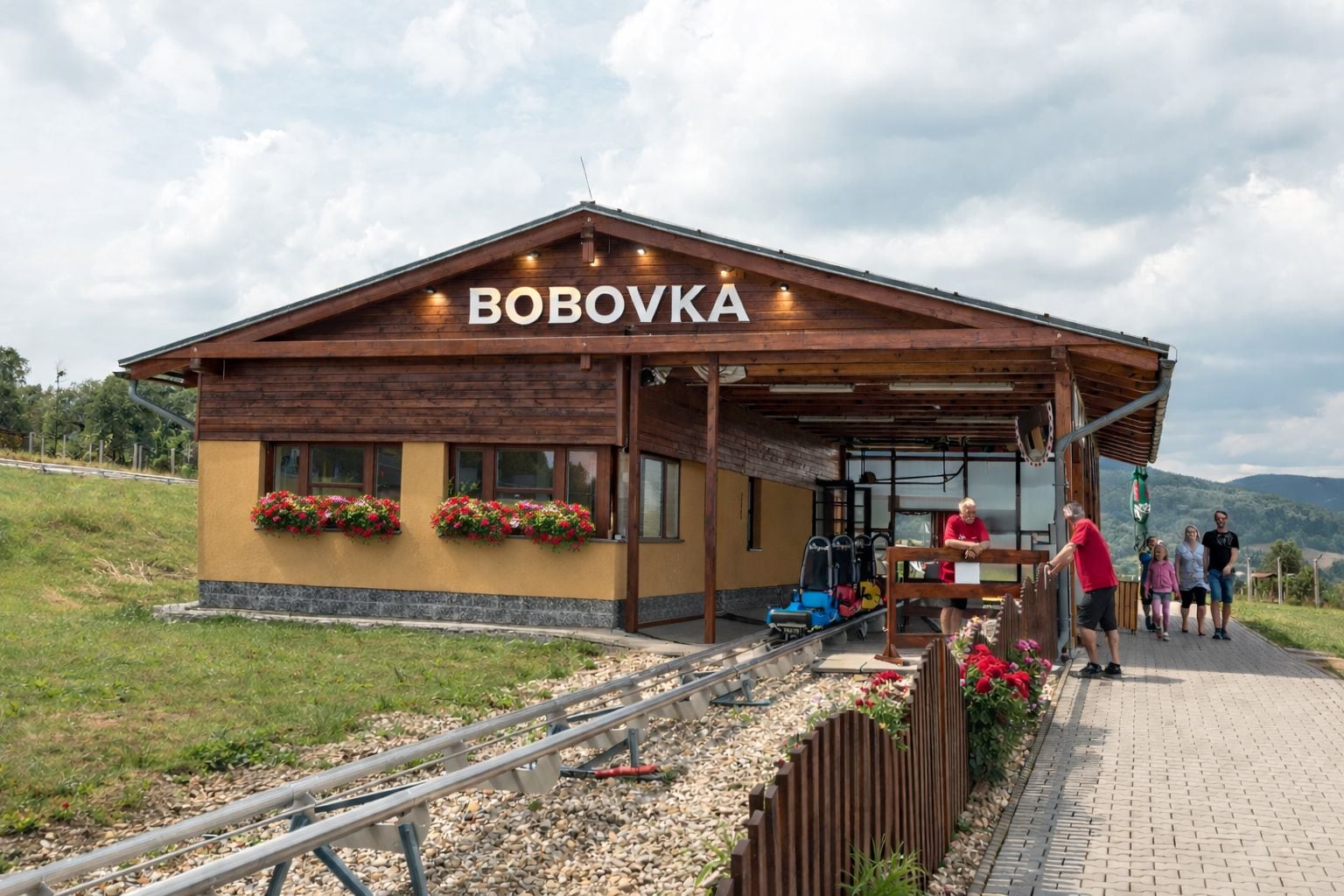 Ein Holzhaus mit der Aufschrift 'BOBOVKA' an der Fassade, ein überdachter Außenbereich mit Sitzgelegenheiten und ein Kiesweg, der zum Eingang führt.