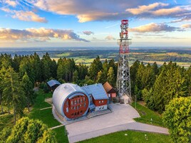 A modern building with a unique curved roof, surrounded by a forested area and a tall communication tower.