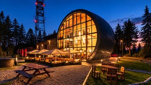 A modern building with a large glass facade, outdoor seating area with umbrellas, and a stone wall. - Hotel Obří Sud (Dlouhý Most)