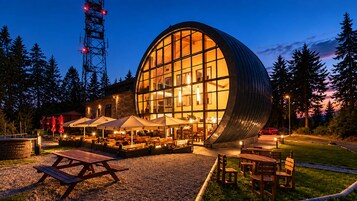 A modern building with a large glass facade, outdoor seating area with umbrellas, and a stone wall.