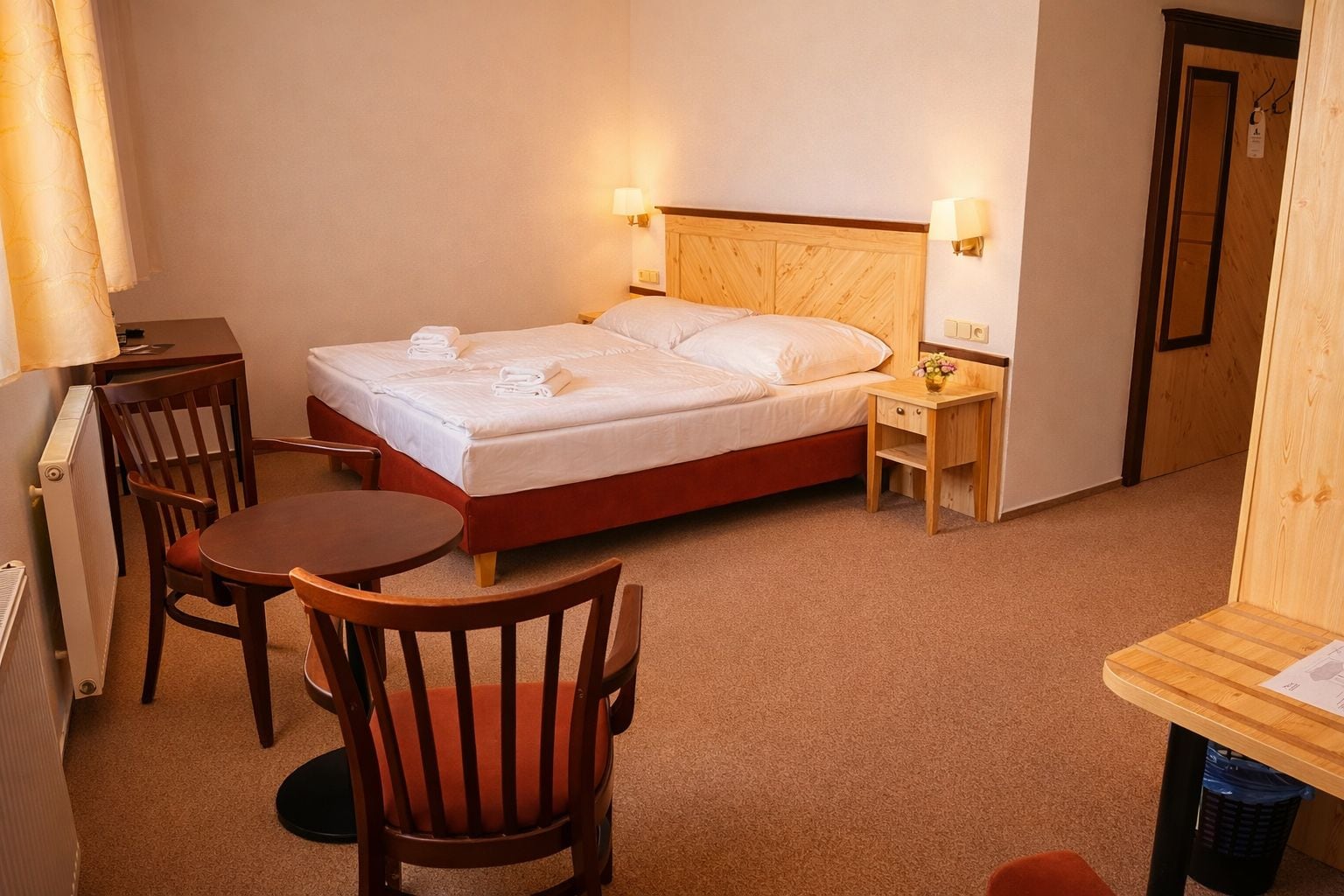 Habitación de hotel con cama, mesita de noche, una silla, una pequeña mesa y un radiador.