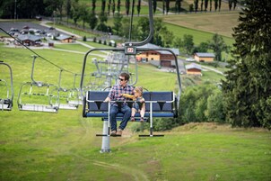 Una persona y un niño en un asiento del telesilla con un letrero con el número 22, rodeados de vegetación y edificios al fondo.