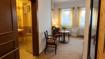 Habitación de hotel con puerta de madera, una pequeña zona de comedor con mesa y sillas, ventana con cortinas y un radiador.