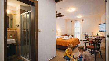 Quarto de hotel com chuveiro em cabine de vidro, cama com colcha laranja, área de refeições com cadeiras e televisão na parede.