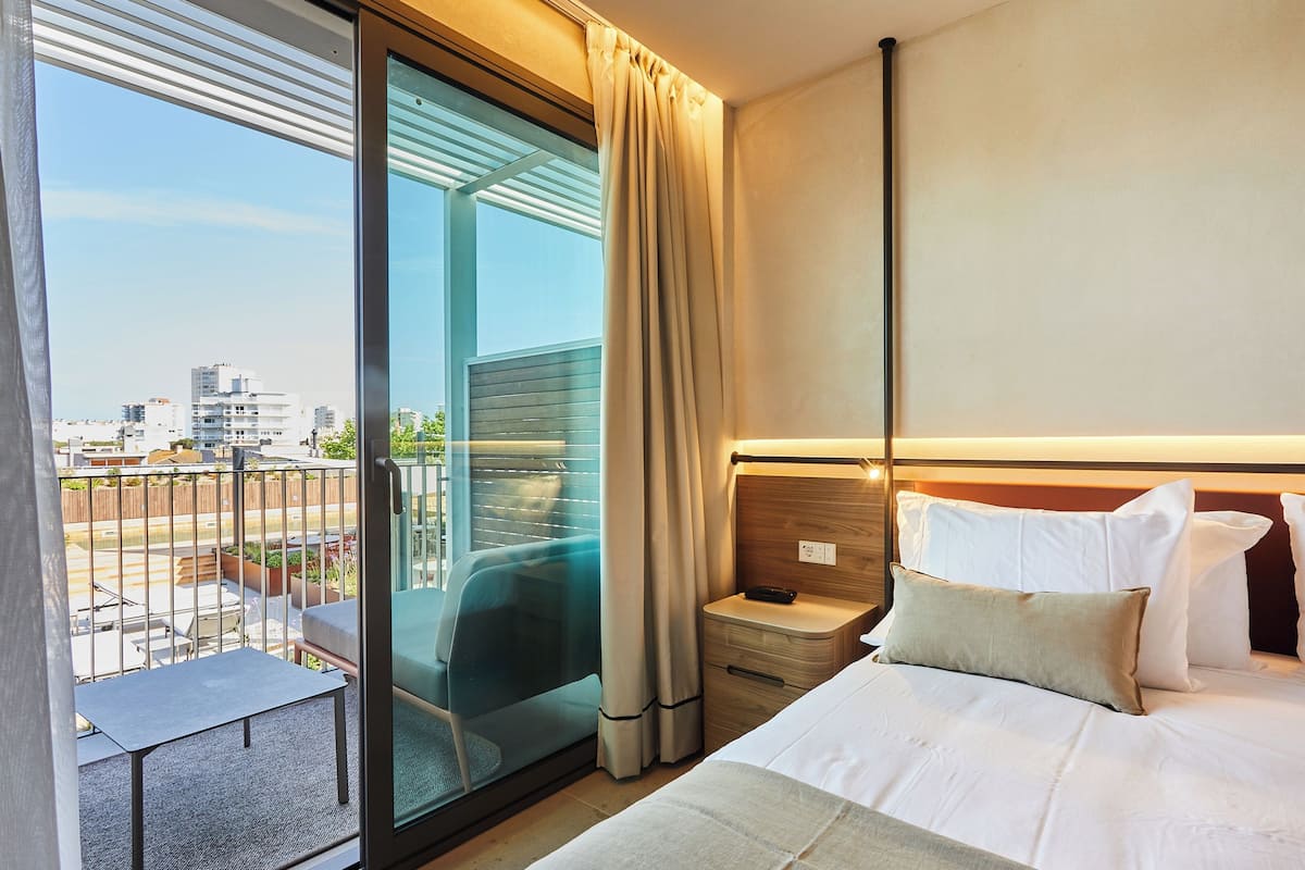 A hotel room with a bed, bedside table, and a view of the city through a glass door.