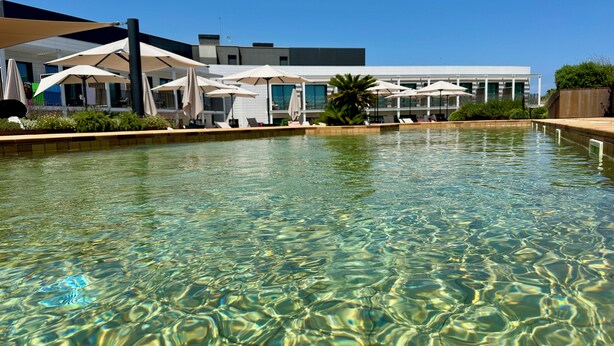 Una piscina cristalina con sillas de descanso y sombrillas, un edificio moderno al fondo, y un cielo azul despejado.