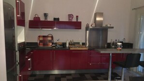 A modern kitchen with red cabinets, a stainless steel range hood, and a bar area with a black chair. - Maison Entièrement Clôturée à 2mn en Voiture de la Plage (Saint-Florent)