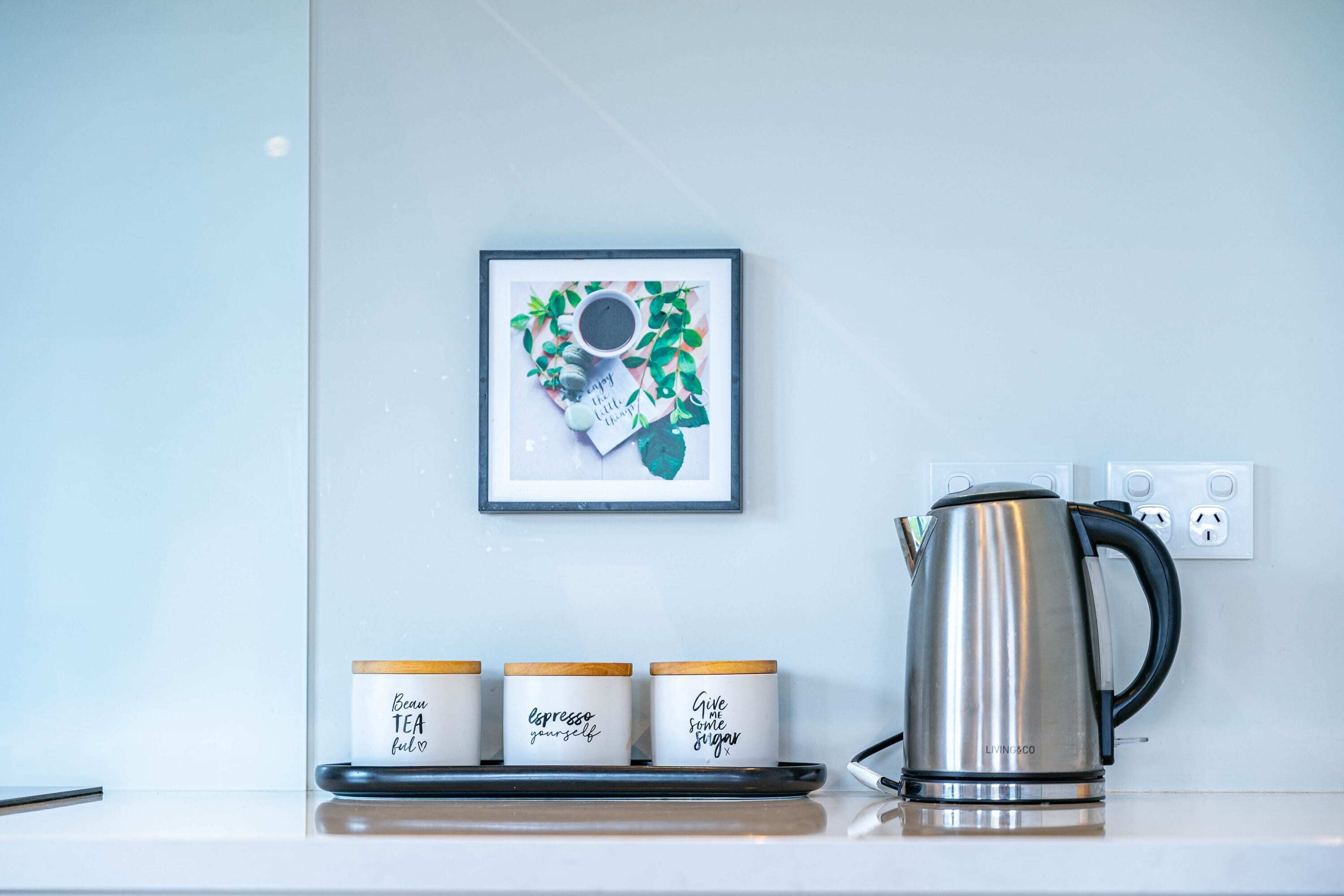 A kitchen counter with a kettle, three jars with labels, and a framed picture on the wall.