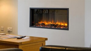 A modern room with a wooden desk, black leather chairs, and a wall-mounted fireplace with visible logs and flames. - Crannog - Three Bedroom Cottage, Sleeps 6 (Llandudno)