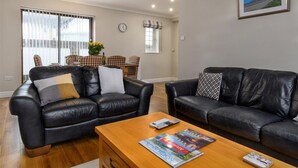 A living room with two black leather sofas, a wooden coffee table, and a dining area with a checkered tablecloth and chairs. - Crannog - Three Bedroom Cottage, Sleeps 6 (Llandudno)