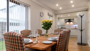 A dining area with a wooden table set for six, plaid chairs, a vase with yellow flowers, and a clock on the wall. - Crannog - Three Bedroom Cottage, Sleeps 6 (Llandudno)