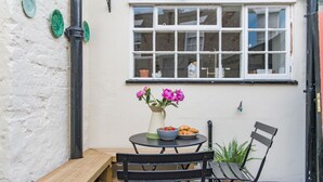 Eine kleine Terrasse mit einer Holzbank, einem Tisch mit Blumen und einem Fenster mit Blick auf die Straße.