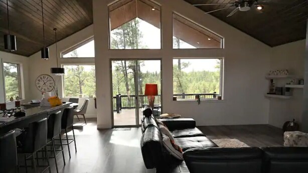 A spacious living area with a long black leather sofa, a kitchen bar with stools, and large windows offering a view of the outdoors. - Mustang Mountain Retreat in Bison Ridge! (Show Low)