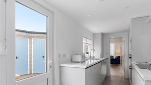 A modern kitchen with white cabinetry, a central island, and a view of a blue door outside. - 4 Bedroom Seaside House, Beach 220m with Free Parking (Sidmouth)