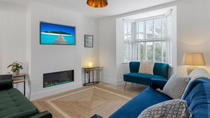 A modern living room with a fireplace, a blue sofa, a teal armchair, and a large window with blinds. - 4 Bedroom Seaside House, Beach 220m with Free Parking (Sidmouth)