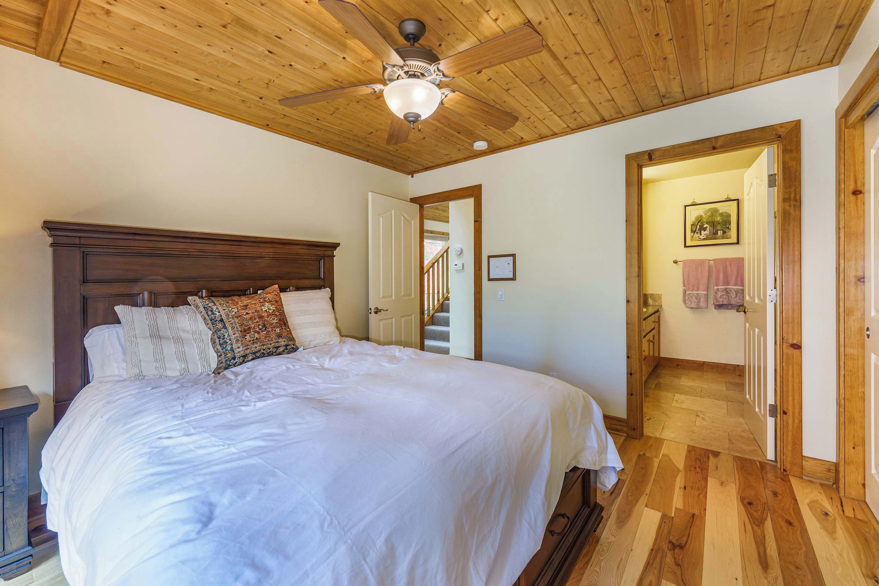 Ein Schlafzimmer mit Deckenventilator aus Holz, einem großen Bett mit weißer Bettwäsche, einem Holzkopfstück und Blick auf einen Flur, der zu einem Badezimmer führt.