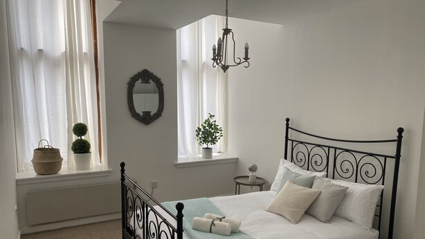 Ein Schlafzimmer mit einem Bett, einem Fenster mit Vorhängen, einem Spiegel, einem kleinen Tisch und einer Topfpflanze.