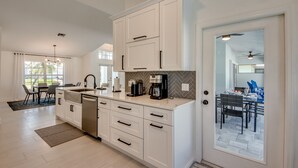Una cucina moderna con mobili bianchi, lavastoviglie e macchina da caffè. Si intravede un'area pranzo attraverso una porta aperta.