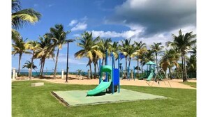 Un terrain de jeux avec un toboggan vert et des balançoires, entouré de palmiers et surplombé d’un ciel dégagé.