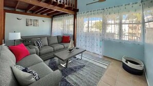 Un salon avec un canapé, une table basse et un panier pour animal de compagnie. Il y a une fenêtre avec des rideaux et une vue sur l’extérieur.