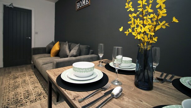 A dining area with a wooden table set for two, a vase with yellow flowers, and a dark wall with a "HOME" sign.