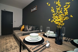 A dining area with a wooden table set for two, a vase with yellow flowers, and a dark wall with a "HOME" sign. - Skyline Remarkable 2-bed Apartment in Swansea (Swansea)