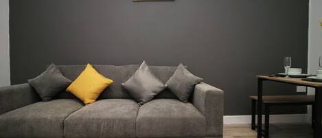 A grey sofa with multiple cushions, a small table with a glass, and a sign on the wall.