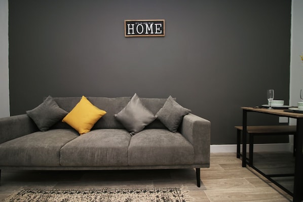 A grey sofa with multiple cushions, a small table with a glass, and a sign on the wall.