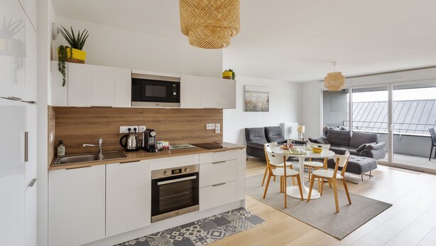 A modern kitchen with white cabinets, a wooden countertop, and a built-in microwave. There is a dining area with a table and chairs, and a living room with a sofa and a large window. - Le Rochenoire - Appt proche plage (Dinard)
