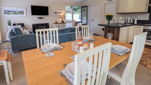 A dining area with a wooden table set for four, white chairs, and a television in the background. - Hill Top Lodge (Windermere)