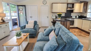 A modern kitchen with white cabinets, a black countertop, and a stainless steel range hood. There is a dining area with a blue sofa and a wooden coffee table. - Hill Top Lodge (Windermere)