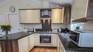 A modern kitchen with a central island, built-in oven, microwave, and a clock on the wall. - Hill Top Lodge (Windermere)